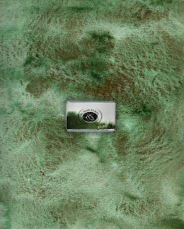 A full pan of December 2025, a new paint by Roman Szmal Aquarius, sits in front of a background made of a swatch of this paint. The paint is green with copious flecks of earthy brown.