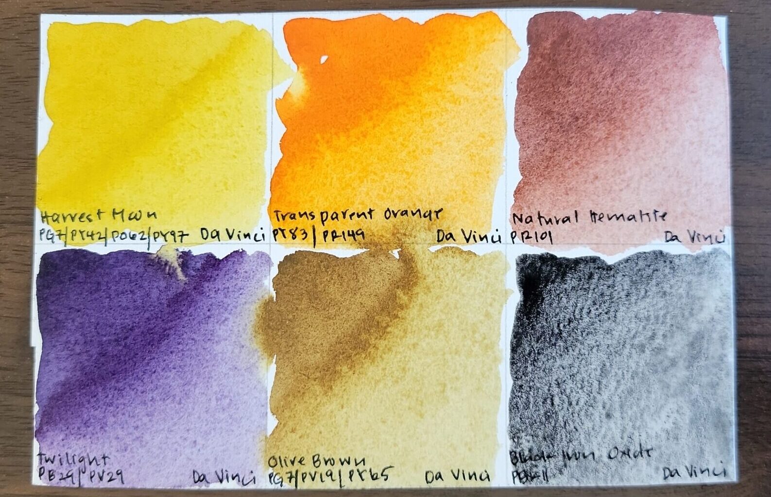 Swatches of the six paints in the Harvest Set by Da Vinci - a yellow, orange, red-brown, violet, earthy yellow, and black hue - sit atop a wood table.